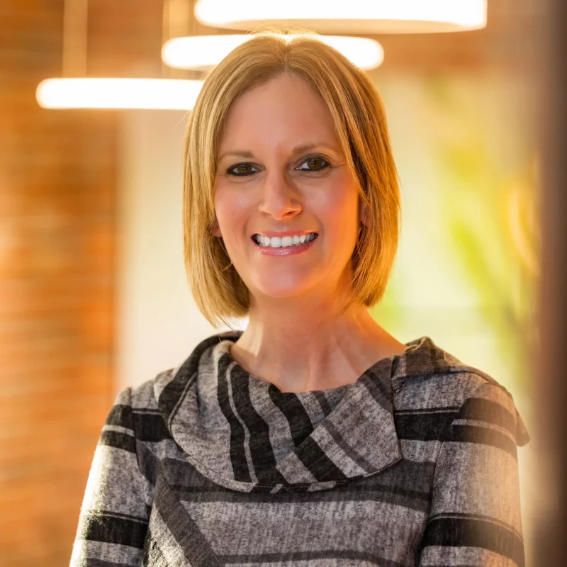 Smiling woman, Jessica Gillespie, with short blond hair and a gray patterned top, stands indoors under warm lighting.