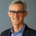 Smiling man with gray hair and glasses, resembling Julien Smith, wearing a checked shirt and dark blazer, against a gray background.