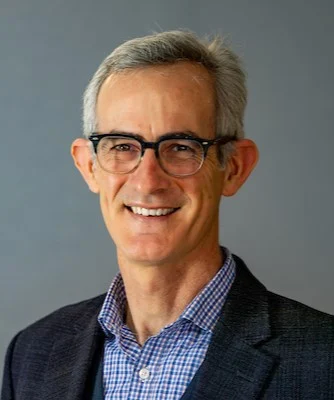 Smiling man with gray hair and glasses, resembling Julien Smith, wearing a checked shirt and dark blazer, against a gray background.
