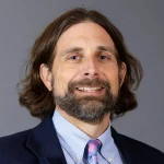 Douglas Wassilak, a man with shoulder-length brown hair and a beard, smiles in a suit against a gray background.