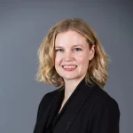 Sara Gundersen, Ph.D., with blonde wavy hair, smiles warmly in a black blazer against a plain grey background.