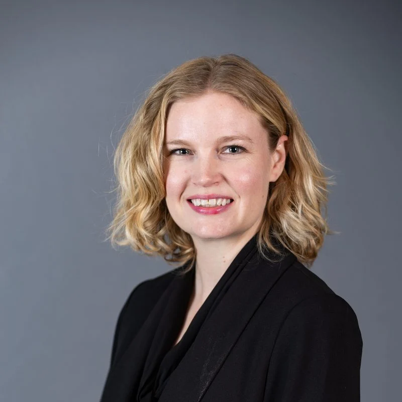 Sara Gundersen, Ph.D., with blonde wavy hair, smiles warmly in a black blazer against a plain grey background.