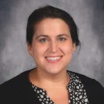 Christine Roberts, a woman with dark hair, smiles while wearing a black and white polka dot blouse and a black sweater.