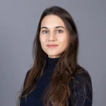 Flavia Mendonca, PhD, with long brown hair, wearing a dark top, poses against a plain gray background.