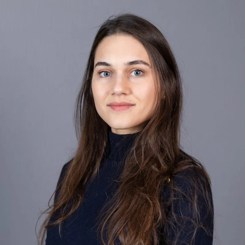 Flavia Mendonca, PhD, with long brown hair, wearing a dark top, poses against a plain gray background.