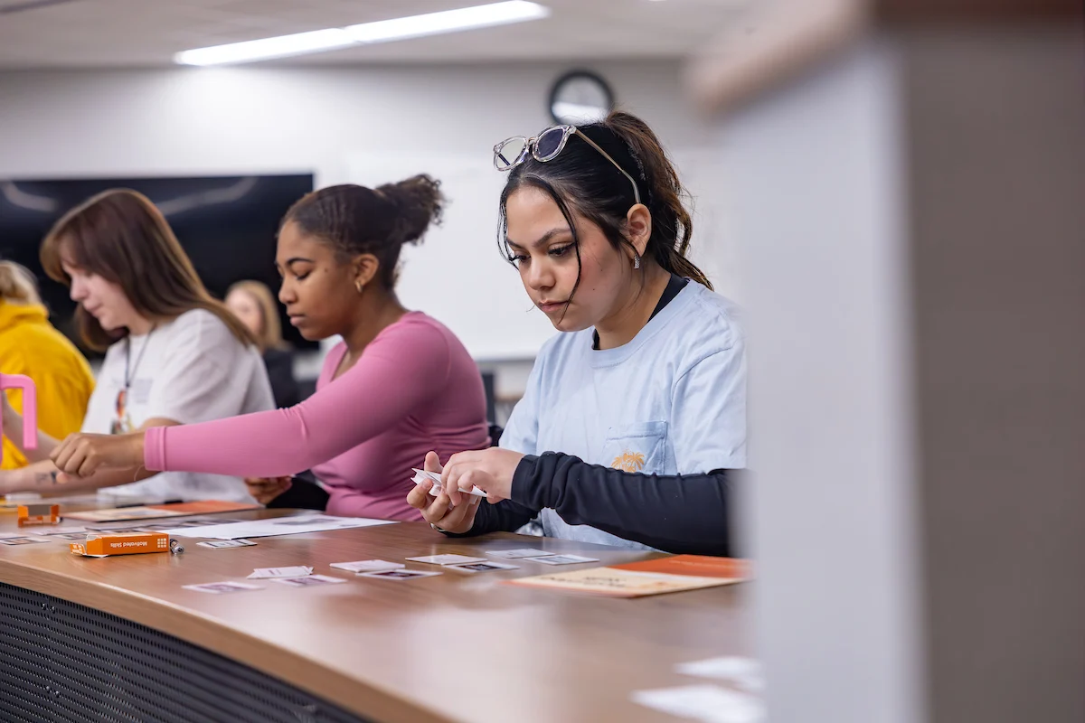 Three young women sit at a desk, focused on organizing papers and cards in a classroom setting.
