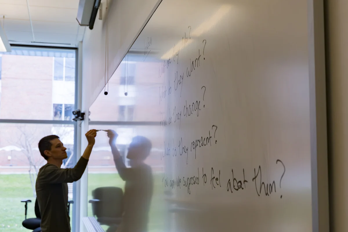 Person writing questions on a large whiteboard in a classroom with windows showing a grassy outdoor area, preparing material related to Academic Policies.