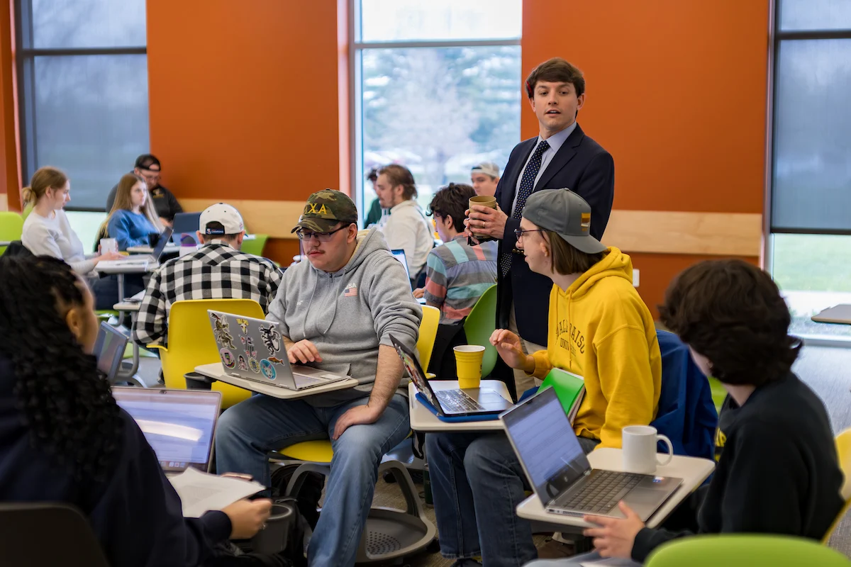 A teacher stands among students working on laptops in a bright, modern classroom, guiding them through important academic policies.