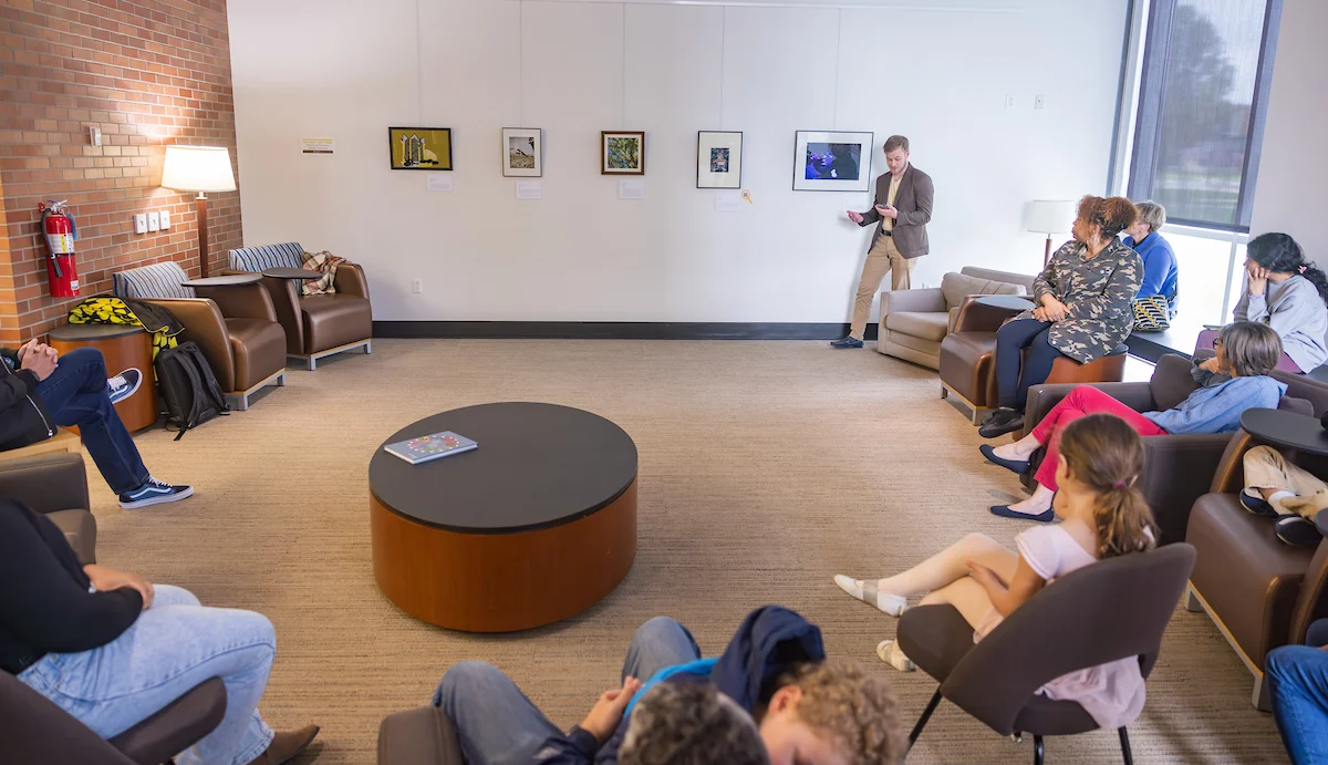 A group of people sit in chairs while a man stands and presents art on a wall in a gallery room, showcasing the gallery’s curated services.