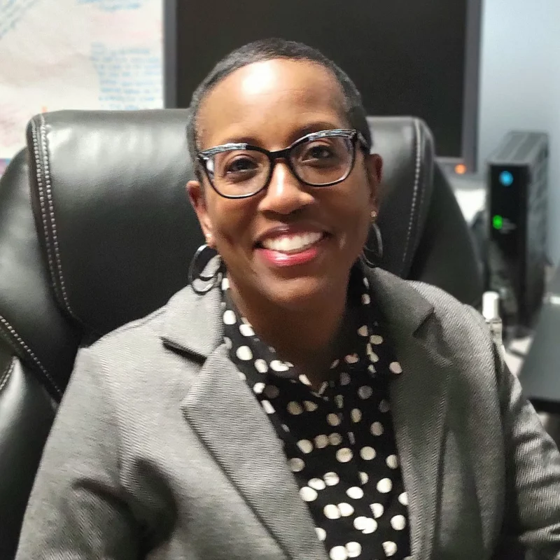 Barbara Dahl, a smiling woman with glasses and a polka dot shirt, sits in a black office chair at her desk.