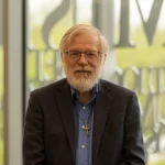 James Nelson, Ph.D., an older man with glasses, a beard, and a cross necklace, wearing a blue shirt and blazer, smiles indoors.