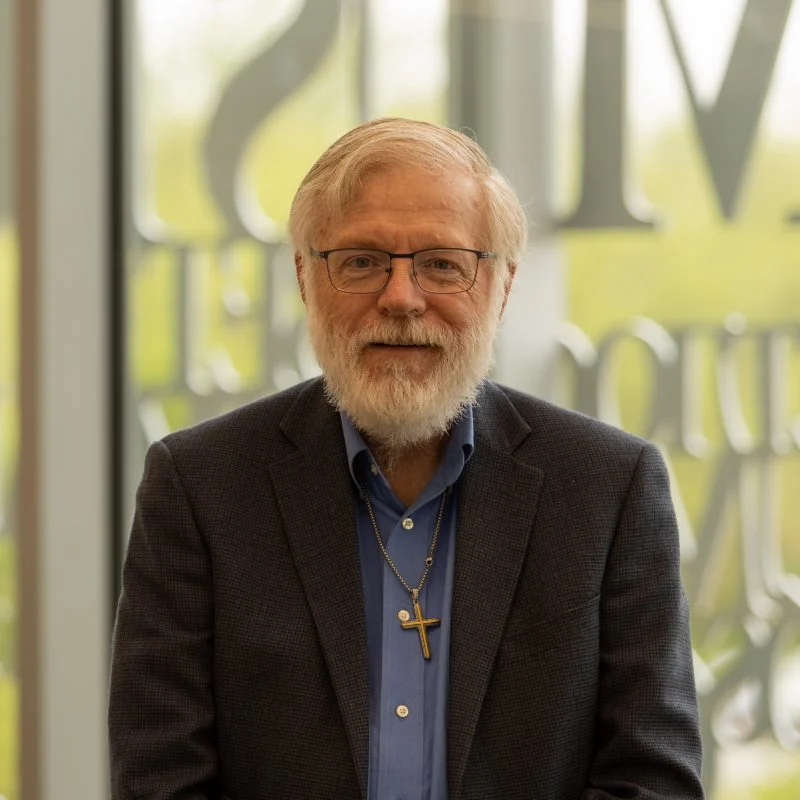 James Nelson, Ph.D., an older man with glasses, a beard, and a cross necklace, wearing a blue shirt and blazer, smiles indoors.