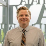 Smiling man, Blake Bent, Ph.D., in a light shirt and patterned tie stands indoors with large letters on the window behind him.