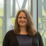Jennifer Winquist, Ph.D., with long brown hair, smiles as she stands indoors by large windows, wearing a dark top and cardigan.