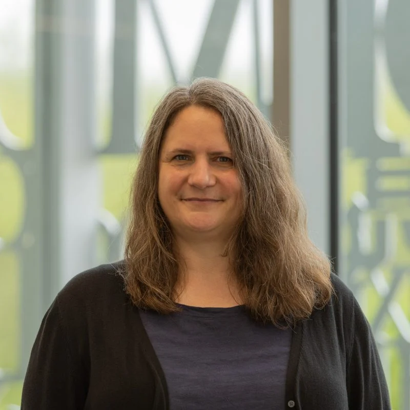Jennifer Winquist, Ph.D., with long brown hair, smiles as she stands indoors by large windows, wearing a dark top and cardigan.