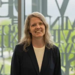 Smiling woman with long, wavy hair wearing a dark blazer, standing indoors in front of a large window—Angela Vernon, Ph.D.