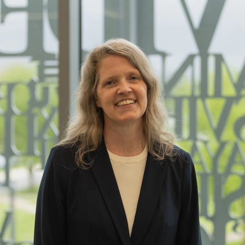 Smiling woman with long, wavy hair wearing a dark blazer, standing indoors in front of a large window—Angela Vernon, Ph.D.