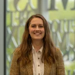 A woman with long brown hair smiles, wearing a plaid blazer and white shirt, standing indoors at Holly Cross.