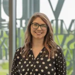 Ellen Ratajack, Ph.D., a woman with glasses and long wavy hair, wears a black polka-dot shirt and smiles indoors.
