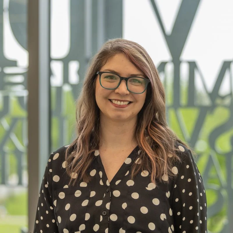Ellen Ratajack, Ph.D., a woman with glasses and long wavy hair, wears a black polka-dot shirt and smiles indoors.