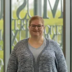 Suzy Wise, LPC, NCC, with short hair and glasses, smiles while standing indoors in front of large glass windows.