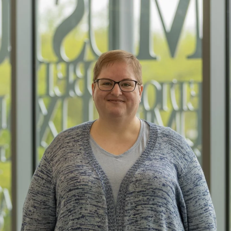 Suzy Wise, LPC, NCC, with short hair and glasses, smiles while standing indoors in front of large glass windows.