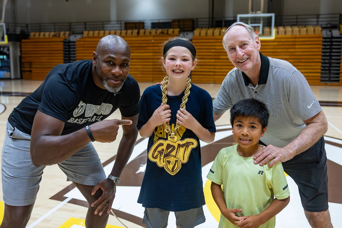 Two men and two kids smile on a basketball court; one kid, sporting a big "GRIT" necklace and headband, shows the power of teamwork—perfect for Marketing and Communications inspiration.