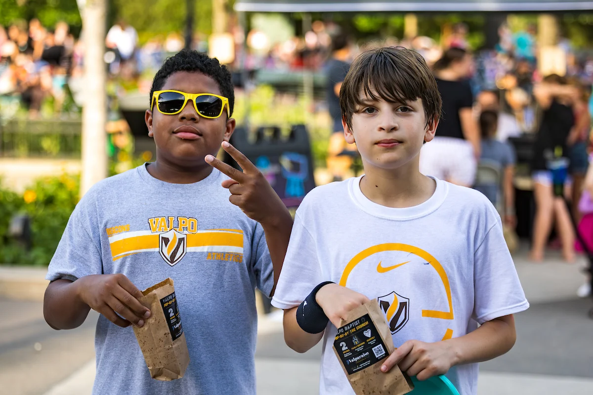 Two boys in Valparaiso T-shirts stand outdoors, holding snack bags; one flashes a peace sign and wears sunglasses, capturing a perfect moment for Marketing and Communications.