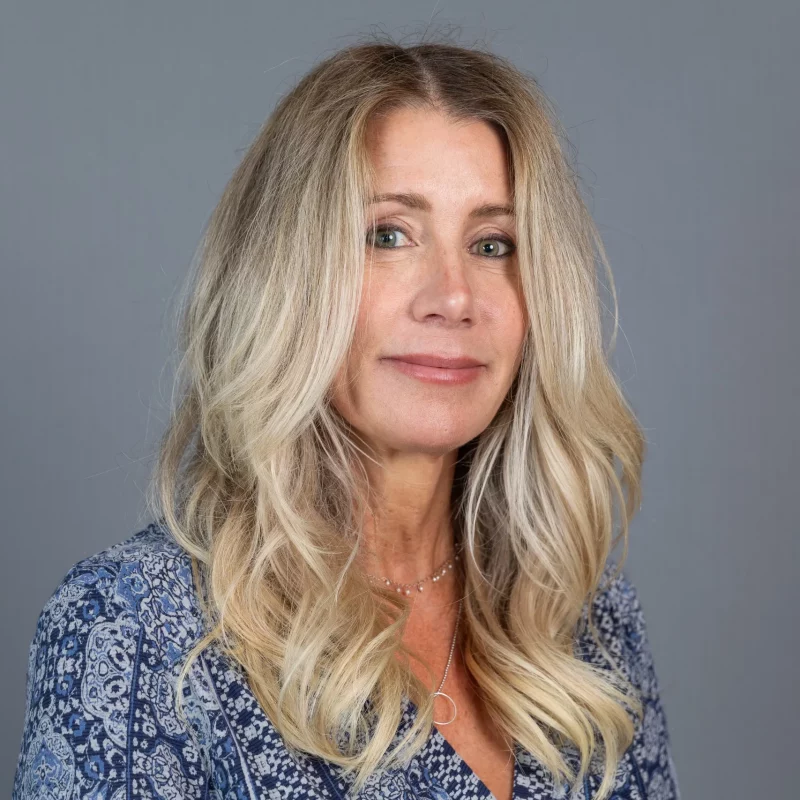 Elizabeth Douglas, a woman with long blonde hair wearing a blue patterned top, smiles slightly against a plain gray background.
