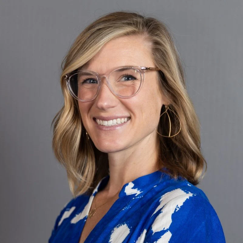Smiling woman with glasses and hoop earrings, wearing a blue and white patterned blouse, on a gray background.