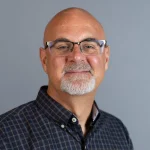 Bald man with glasses, goatee, and checked shirt smiling in front of a plain gray background—Matthew Ringenberg.