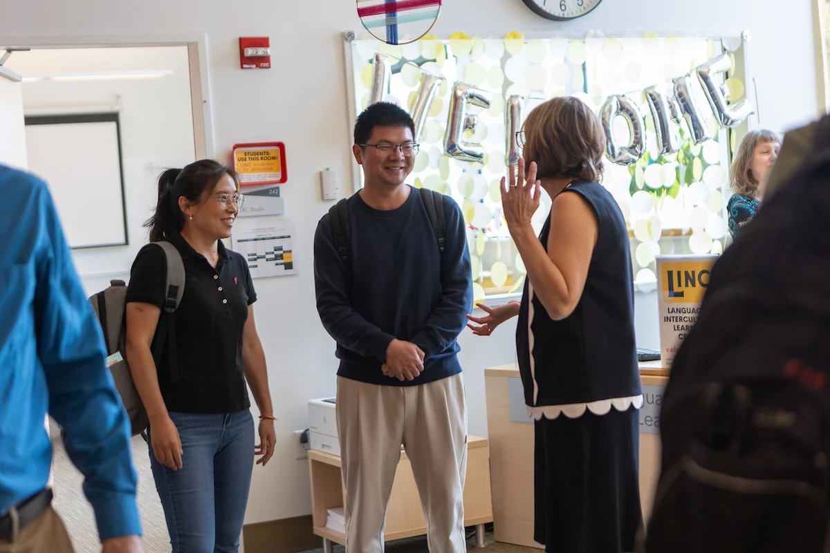 Three people stand and talk happily in a classroom with a "WELCOME" banner in the background, excitedly discussing their upcoming study abroad experiences.