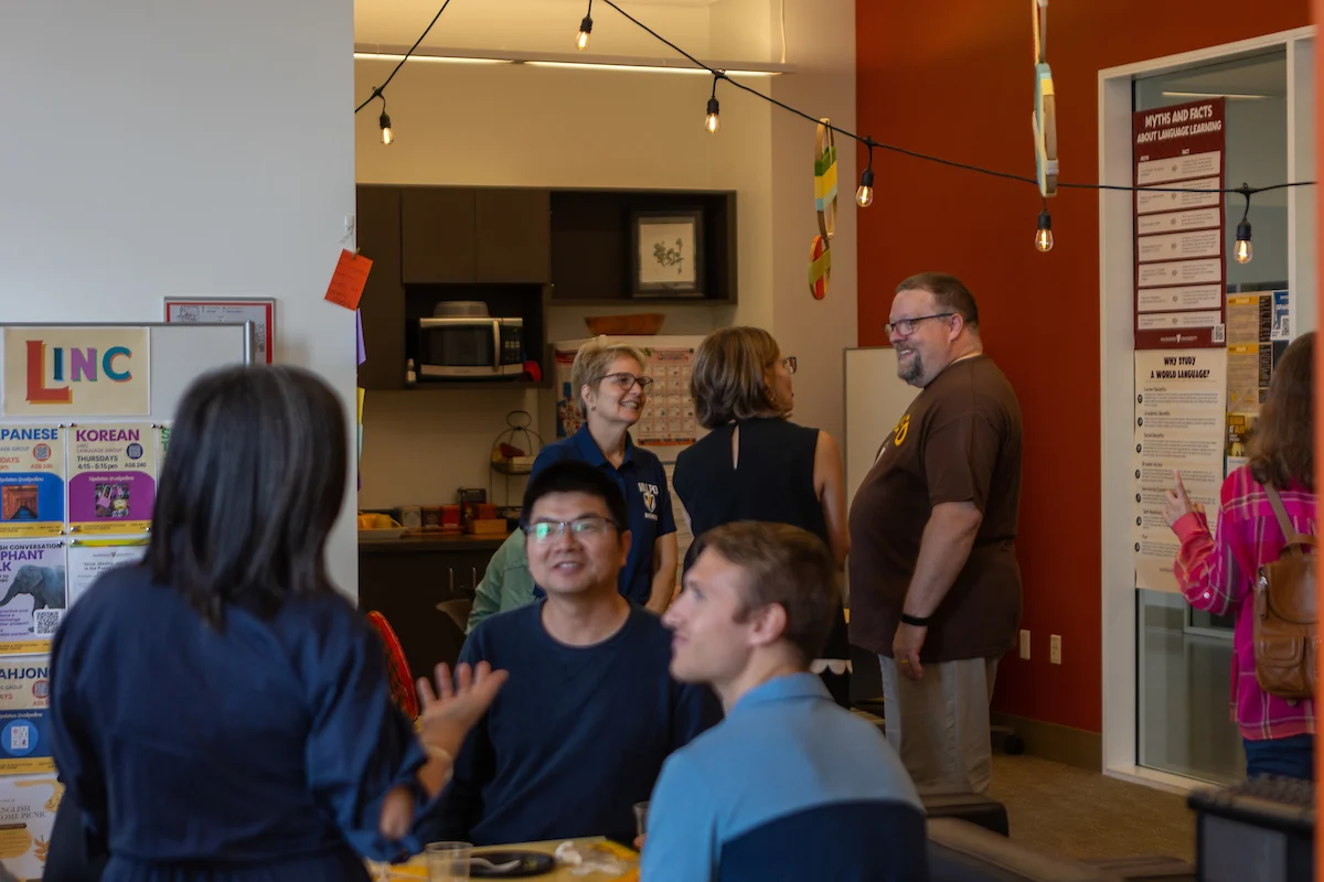 People gather and talk in a brightly lit room decorated with string lights and posters on the wall, sharing their study abroad experiences.