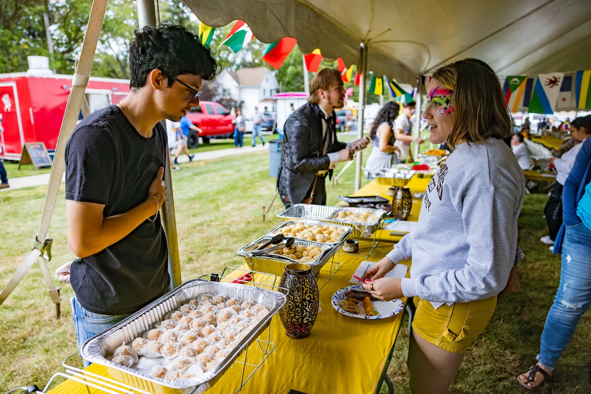 People serving and choosing food at an outdoor festival under a tent, with colorful flags and decorations hosted by International Student Associations.