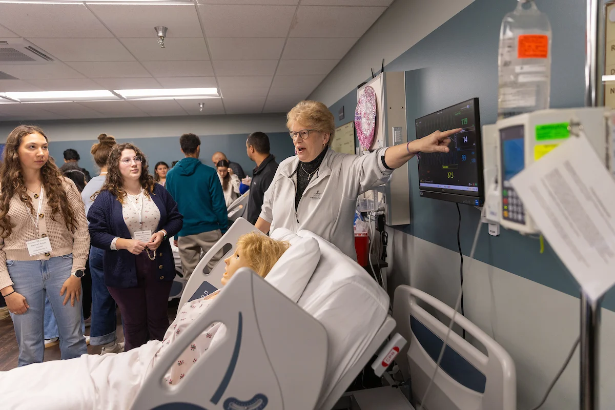 A woman, demonstrating medical assistant techniques, points at a monitor beside a hospital bed with a mannequin, while students observe in a classroom.