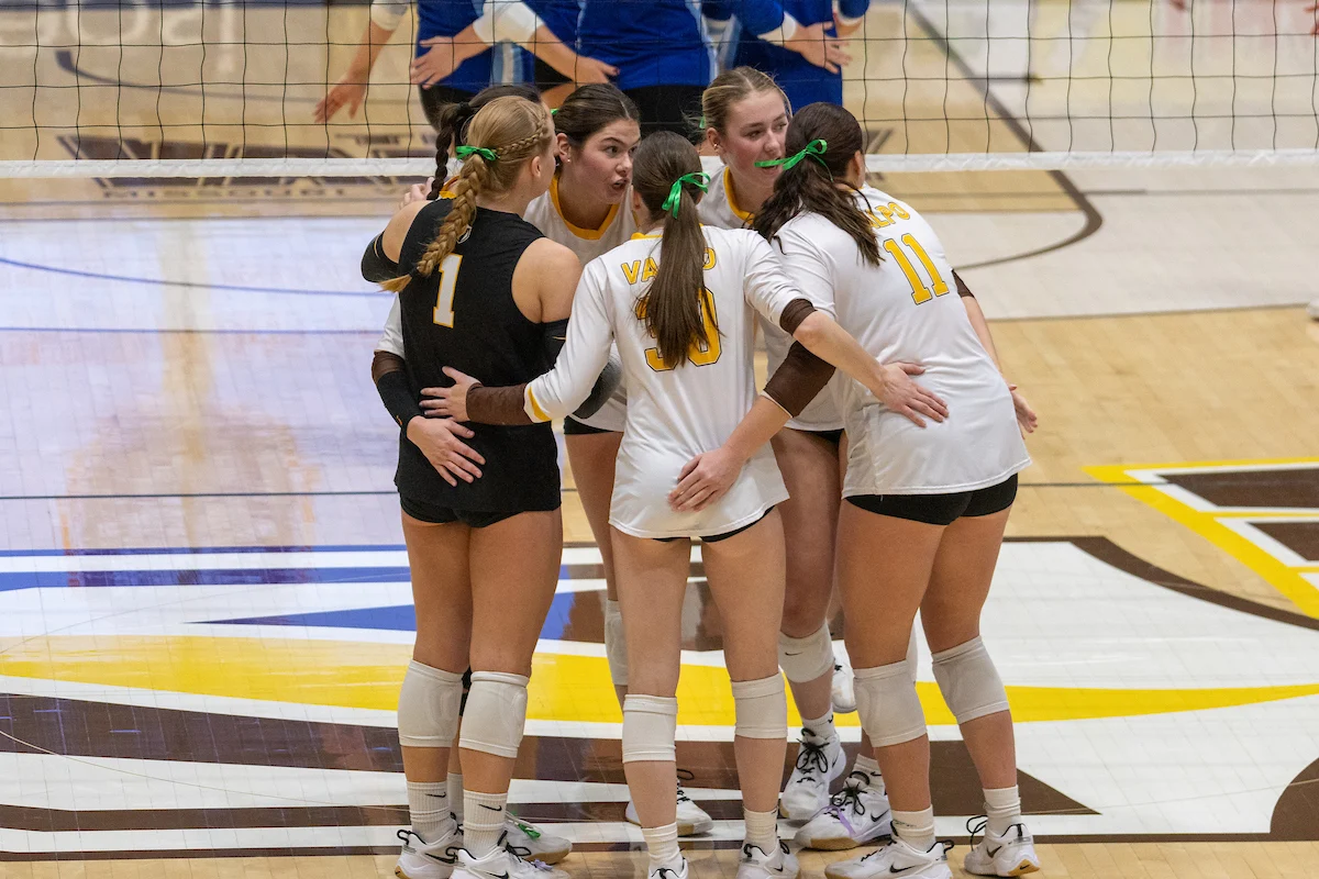 Five female volleyball players in uniforms huddle together on an indoor court, demonstrating teamwork and unity—an inspiring scene for Marketing and Communications efforts.