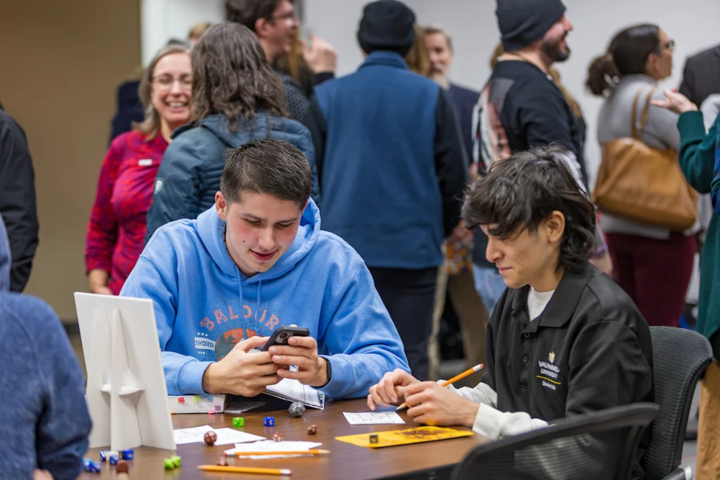 Two people sit at a table playing a game with dice and papers, surrounded by a crowd in the background—an engaging scene at the Academic Success Center.