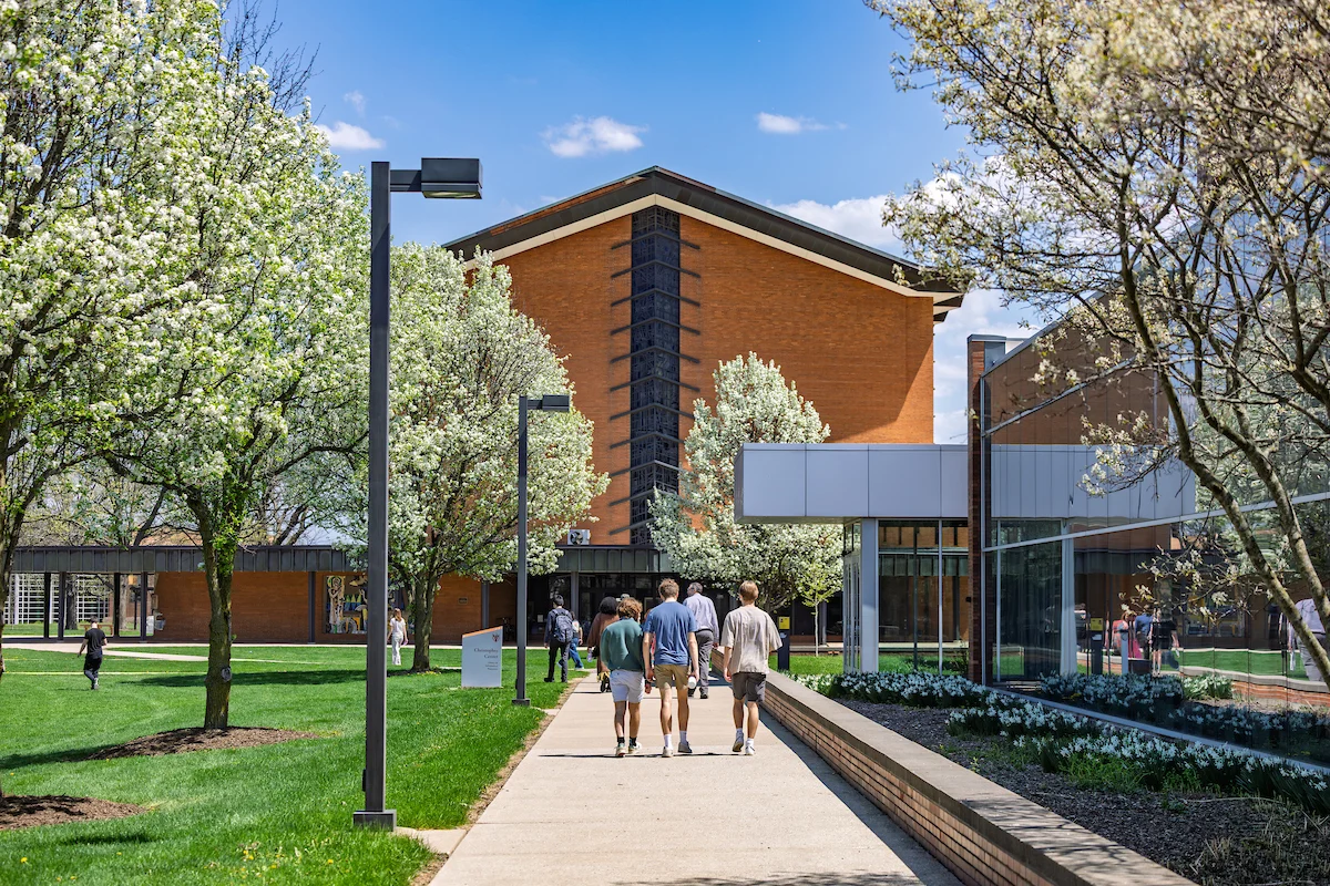 Four people walk along a tree-lined campus path with brick and glass buildings on a sunny day, sharing handouts and discussing helpful how-to’s.