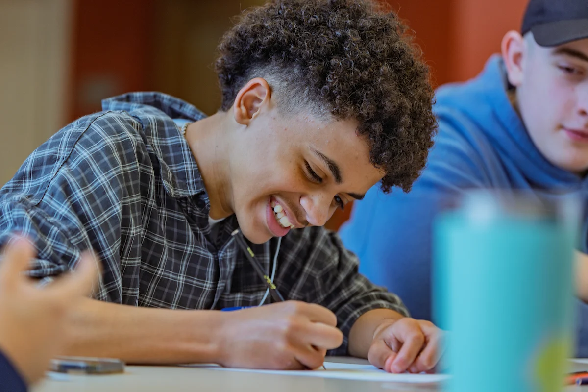 Smiling teen with curly hair writes at a table, sitting next to another student in a blue hoodie as they discuss their study abroad plans.