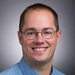 Adam Stepanek, Ph.D., with short brown hair, glasses, and a blue shirt, smiles at the camera against a gray background.