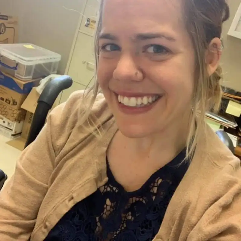 Smiling woman with light brown hair in an office, wearing a tan sweater and navy lace top, representing the friendly staff.