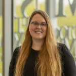 Abbie Thompson, Ph.D., a woman with long blonde hair and glasses, smiles while standing indoors in front of large windows.