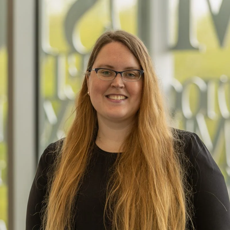 Abbie Thompson, Ph.D., a woman with long blonde hair and glasses, smiles while standing indoors in front of large windows.