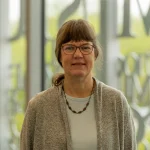 Jeanne Brown, Ph.D., a woman with glasses and brown hair in a ponytail, wearing a grey sweater, stands indoors and smiles.