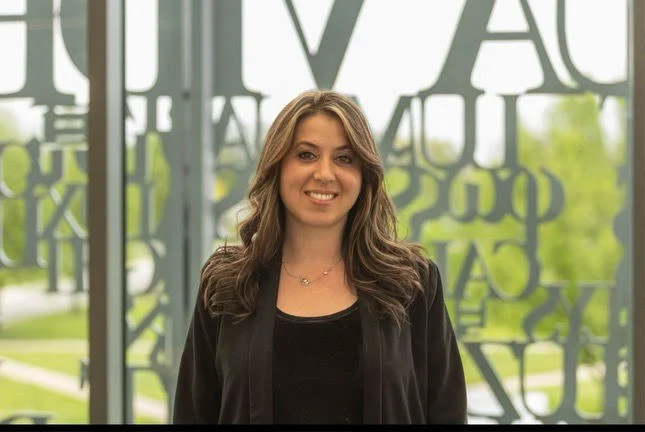 Misty Hatch, NCC, LMHC, smiles warmly, standing indoors in front of large glass windows with letter designs.
