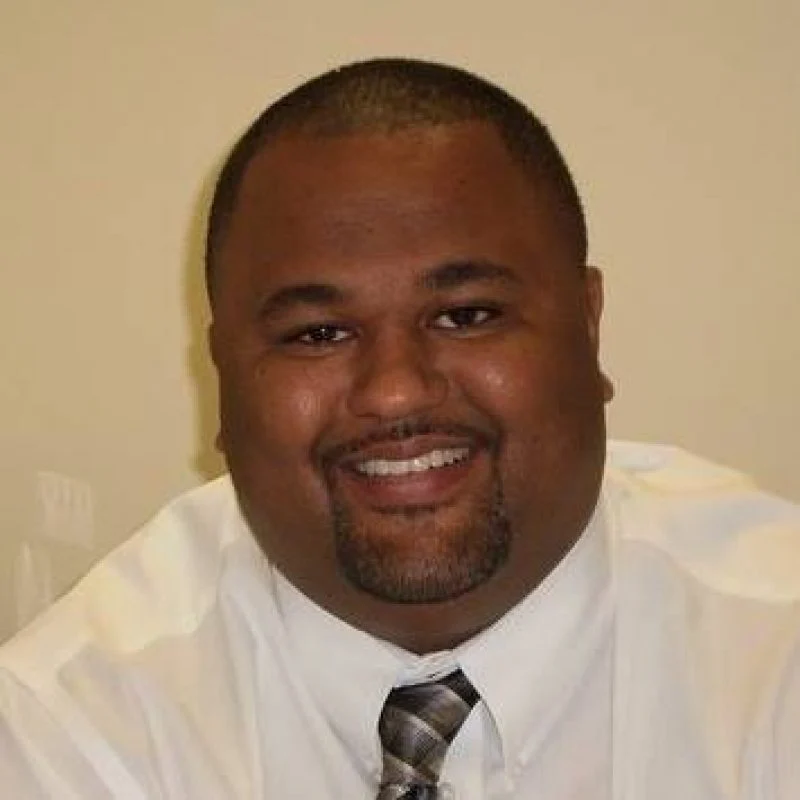 A man in a white shirt and tie smiles at the camera against a plain background.
