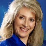 A smiling woman with long blonde hair, Adrienne Sobolak, wearing a blue jacket against a blue background.