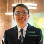 A man in a suit and tie, possibly Jiun-Lin Chen, Ph.D. or Alex Chen, smiles indoors with modern lights and a blurred background behind him.