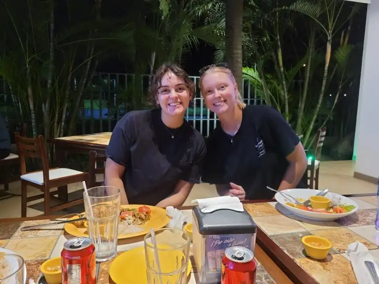 Two women smile at a dinner table outdoors at night, with plates of food and drinks in front of them, as gentle WAVES can be heard in the background.