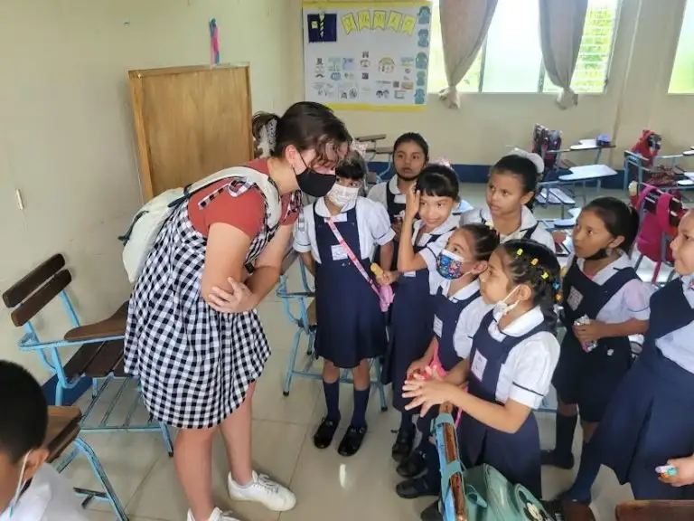 A woman WAVES as she talks to a group of young students in uniforms inside a classroom.